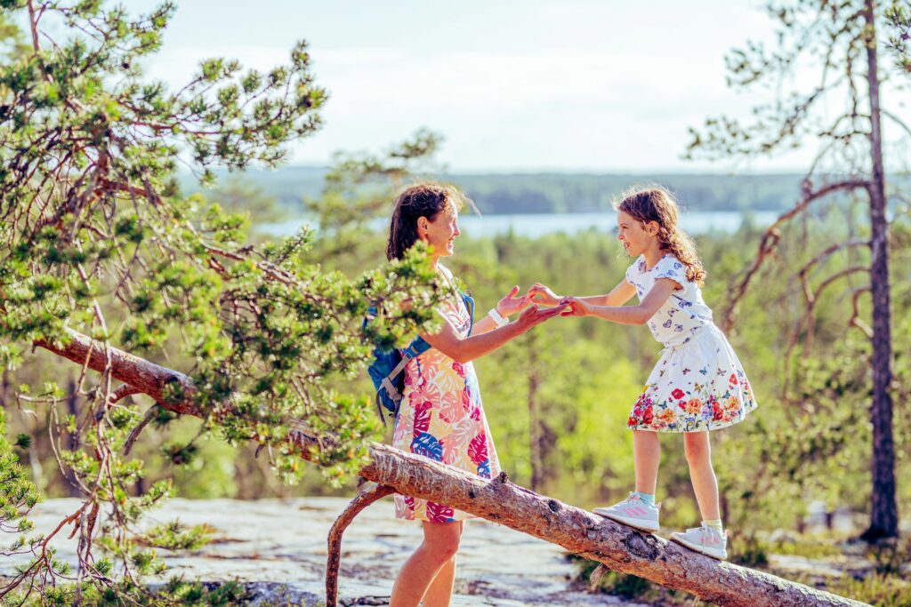 A girl walks along the trunk of a tree while her mother holds her hands.  Behind them is the forest and a lake scene.