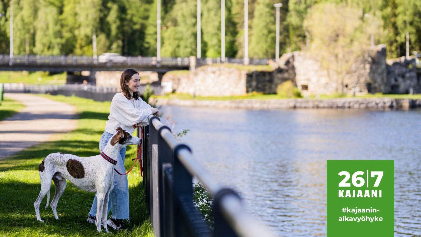 Nainen ja koira katselevat Kajaaninjoelle. Takana näkyy Kajaanin rauniolinna.
