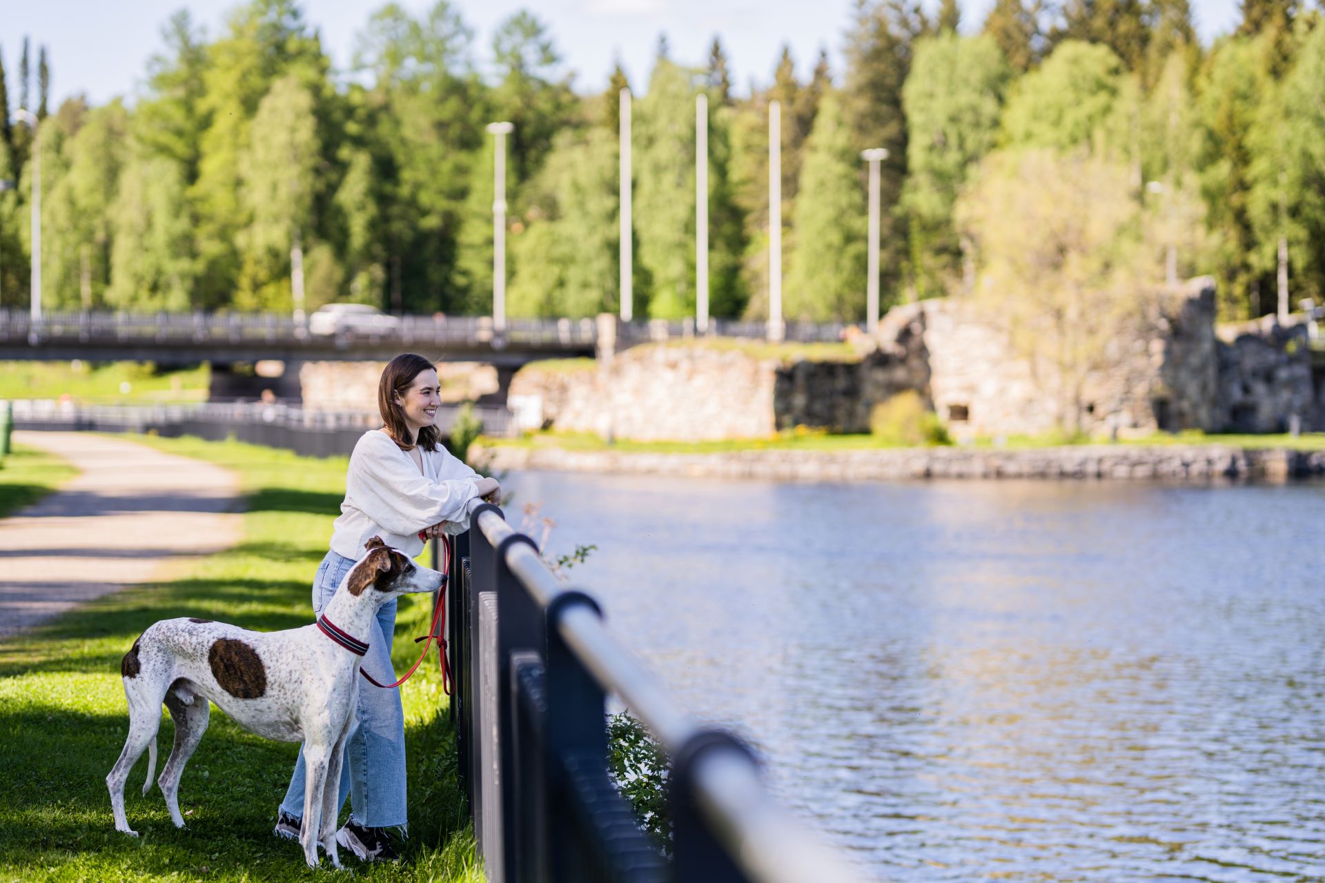 Nainen katselee koiran kanssa Kajaaninjoelle. Takana rauniolinna.
