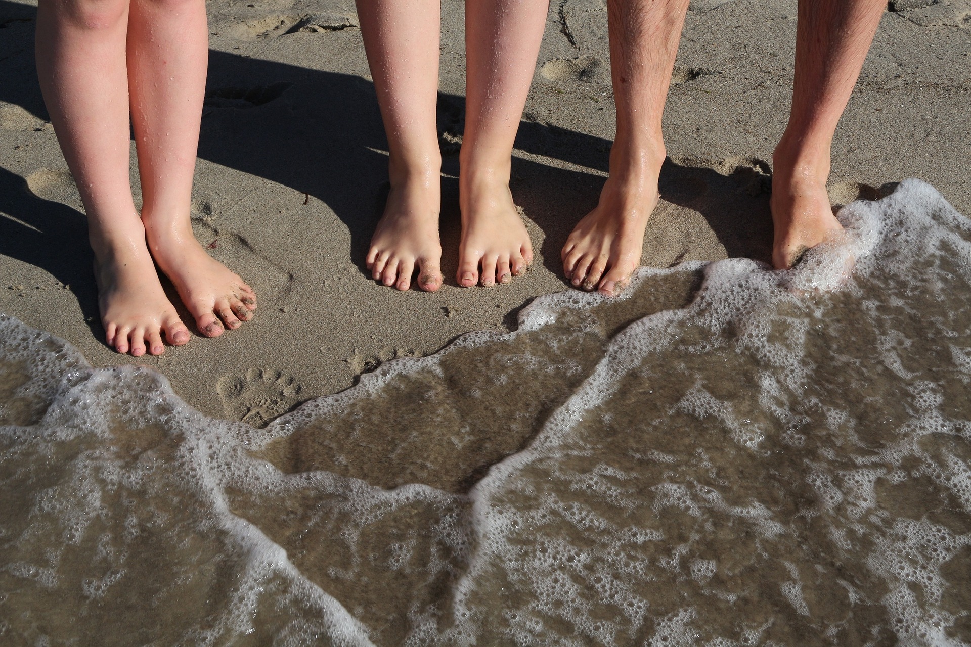Kolme ihmistä paljain jaloin hiekalla rannassa, vain jalat näkyvät ihmisistä