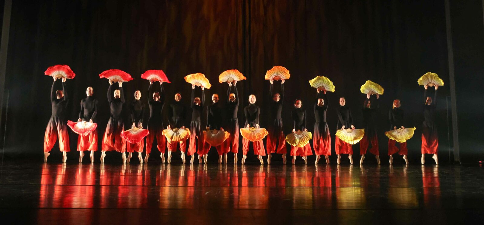 tanssijoita rivissä lavalla mustaa taustaa vasten, oranssit viuhkat kädessä. heillä on mustat paidat ja oranssit housut.