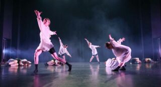 Ryhmä tanssijoita valkoisissa puvuissa, osa makaa lavalla ja osa seisoo. Sumuinen, musta tausta ja kirkkaat valot.