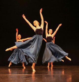Tanssijat seisovat muodostelmassaan toinen jalka ilmassa, sinisissä puvuissaan. Kellertävät valot ja musta tausta.