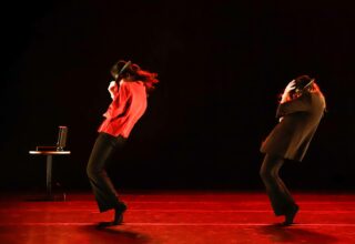 Kaksi tanssijaa mustaa taustaa vasten, punaisessa valossa.